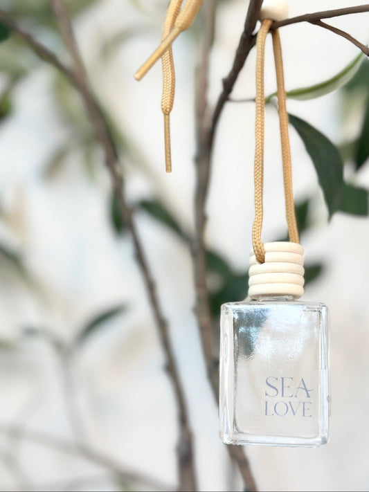 A translucent perfume bottle with the label "sea love" hanging from a beige ribbon on a green leafy branch, with a blurred background suggesting a serene and natural setting.